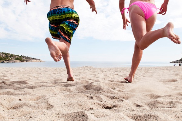 två barn med badkläder, sedda bakifrån, springer mot vattnet på en sandstrand