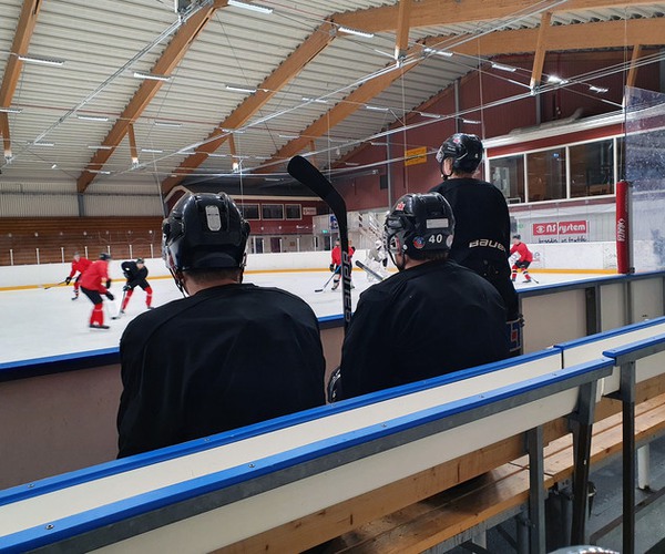 tre ishockeyspelare i ett bås tittar på andra spelare som tränar på isen