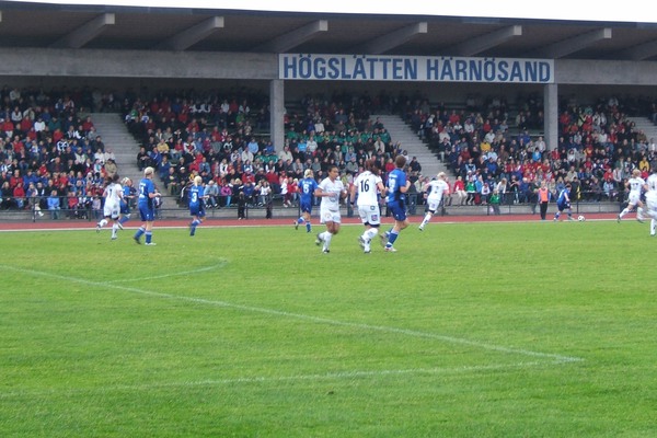 en fotbollsplan där det pågår en match. I bakgrunden en läktare med mycket publik