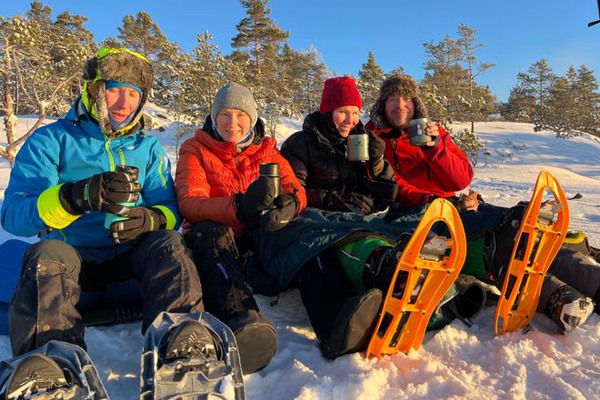 Vänner sitter i snön och fikar tillsammans i solskenet.