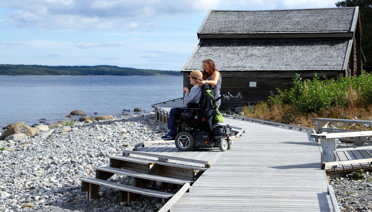 en person sår och en sitter i en rullstol på ett trädäck vid en stenstrand med sjöbodar i bakgrunden