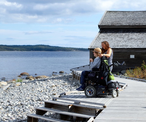 en person sår och en sitter i en rullstol på ett trädäck vid en stenstrand med sjöbodar i bakgrunden